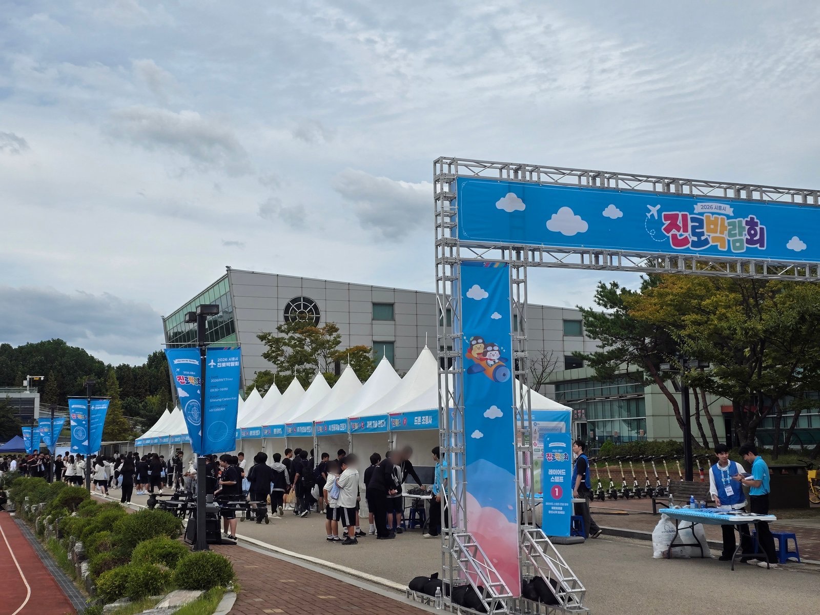 The entrance gate of the 2026 Siheung Career Fair on the GTEC campus, with teenagers filing past the first marquees toward the drone pilot booth