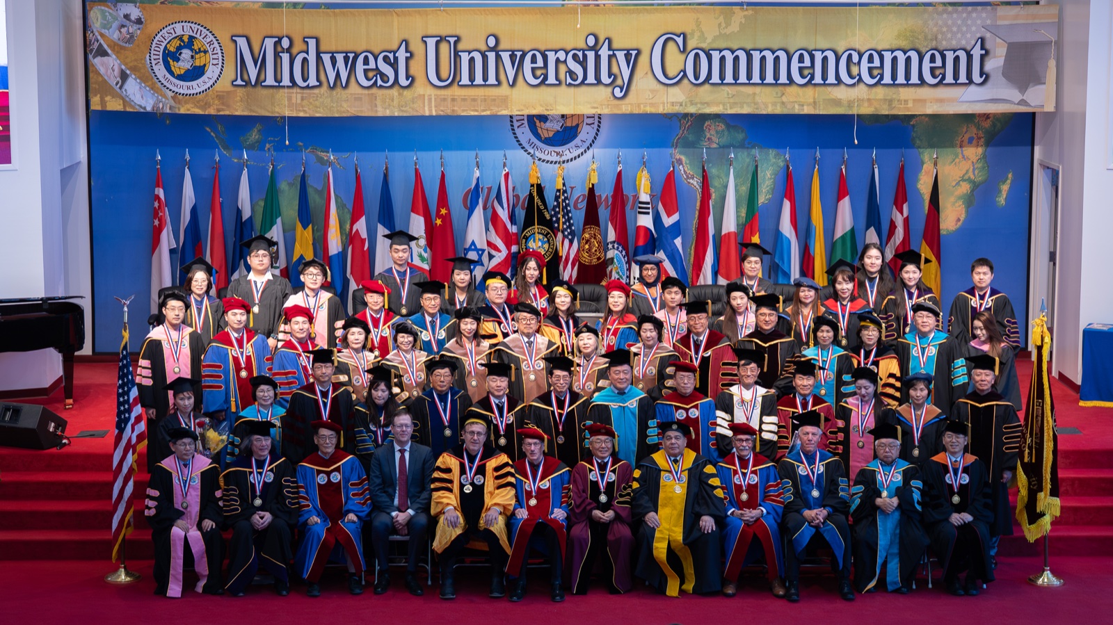 Graduates and faculty of Midwest University on stage at the 2026 commencement, in academic dress before a wall of national flags