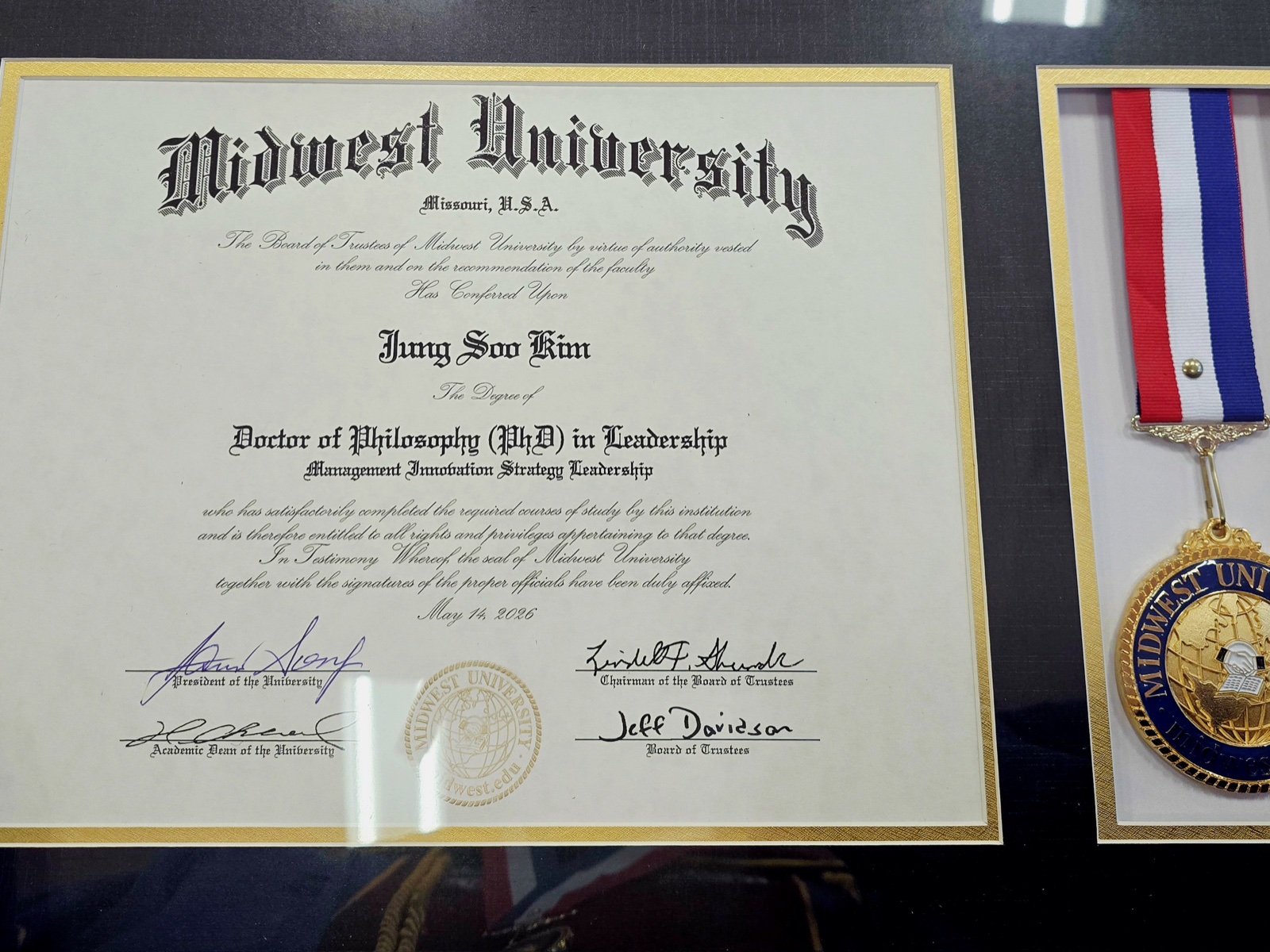 The framed doctoral diploma from Midwest University, dated 14 May 2026, displayed with the university medal on its red, white, and blue ribbon