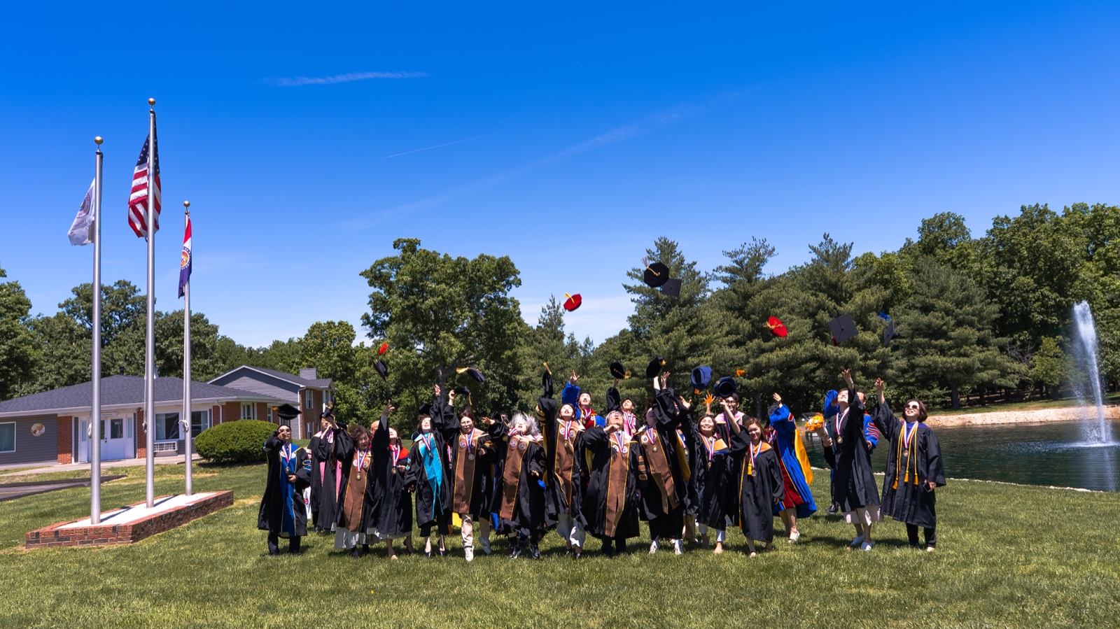 Graduates tossing their caps on the lawn outside the university, with the flags of the United States and Missouri and the campus pond behind them